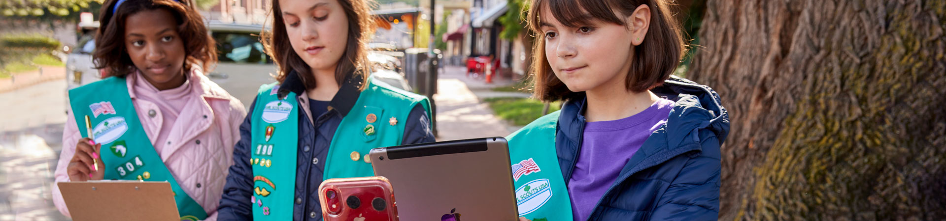 Three Girl Scouts wearing green vests decorated with pins and badges are standing outside. One is holding a clipboard and another is holding an ipad. A hand holds up a red iphone to the Girl Scout with the ipad.  