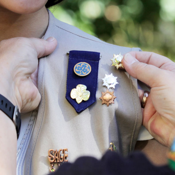 A Girl Scout wearing a khaki-colored vest embellished in a variety of metallic pins stands while an adult adds a gold pin to their vest.