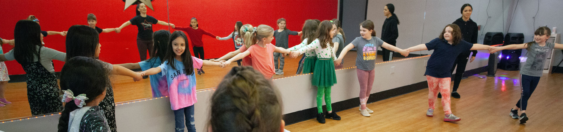  A group of youth Girl Scout members are in a gym with a woode floor and red walls. The Girl Scouts are standing around the perimeter of the room, arms extended, and holding each other's hands.  