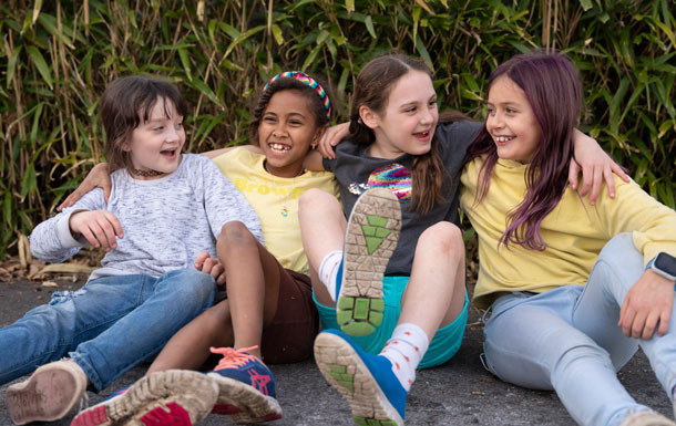 Four Girl Scouts sit on the ground with arms around each other in front of green foliage, looking between each other and smiling.