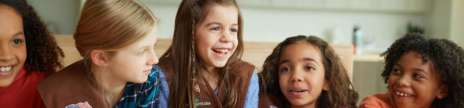  Image Description: Three Girl Scouts wearing brown vests with badges. The Girl Scout in the middle is smiling wide. There is a wooden table in the background. 