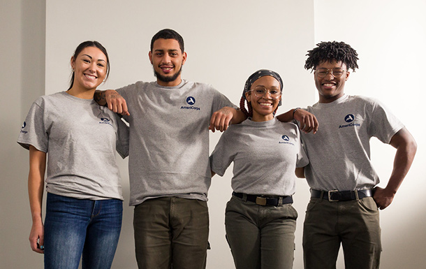 Four AmeriCorps Girl Scout volunteers wearing matching gray shirts stand in a row with their arms around each other's shoulders.