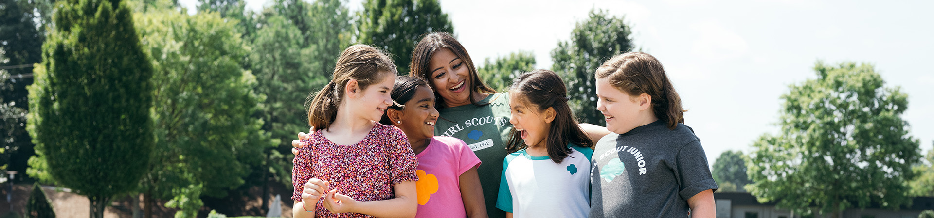 Group of Girl Scouts and an adult volunteer standing outdoors smiling with their arms wrapped around each other. 