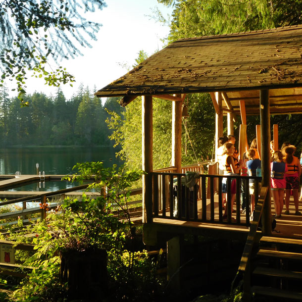 A group of Girl Scouts wearing life jackets and holding oars gather under a covered porch at Girl Scout camp St. Albans. The sun is shining through the trees that surround the lake next to them.