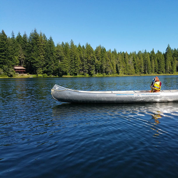 A Girl Scout in a yellow life vest is in a canoe on a blue-watered lake surrounded by evergreen trees at Girl Scout camp.