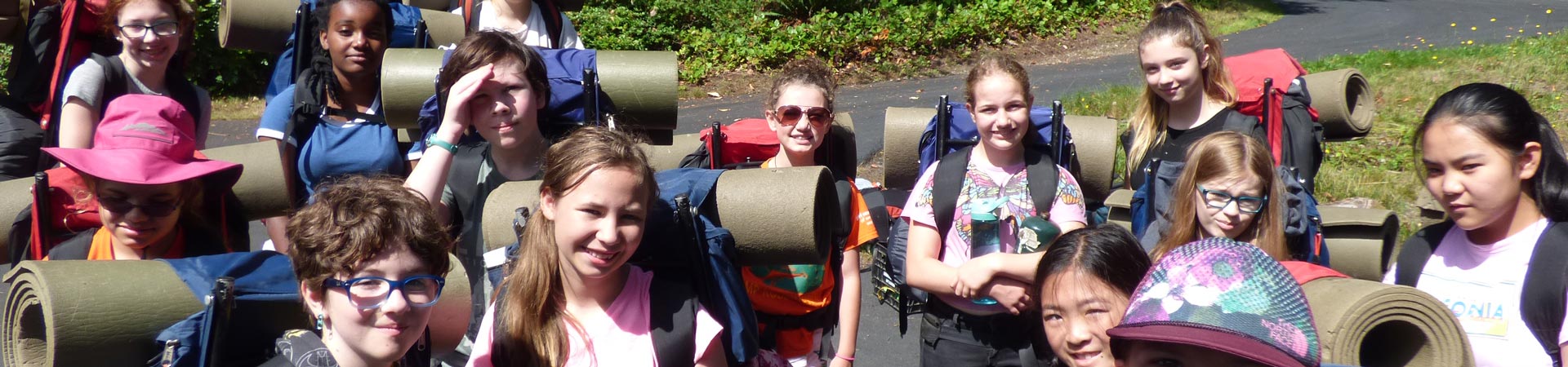  A group of Girl Scouts wearing backpacks standing outdoors near a road at Girl Scout camp. 