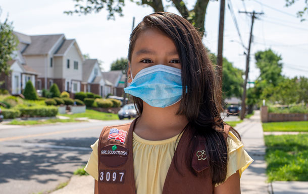 A girl Scout standing on a sidewalk wearing a yellow shirt and brown vest wears a blue face mask.