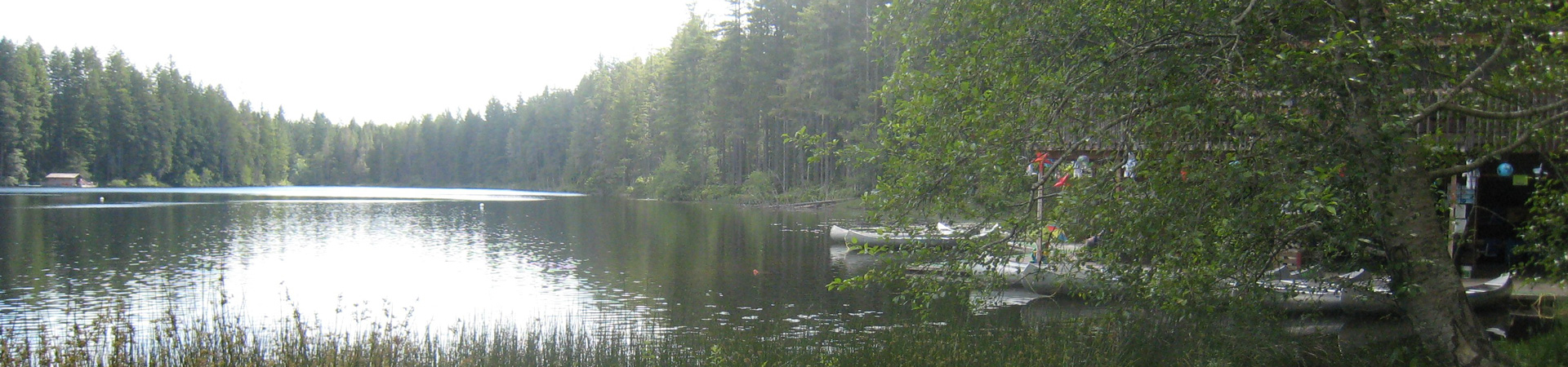  A lake surrounded by trees at Girl Scout camp. Canoes and safety gear are lined up near the shore. 