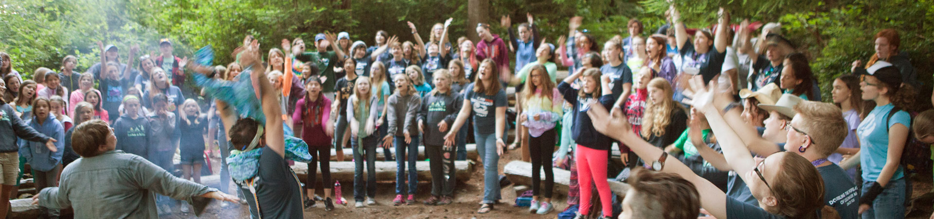  Adult Girl Scout volunteers and staff lead a large group of youth Girl Scout members in song around a campfire. The Girl Scouts are standing near wooden benches with their arms raised. There are trees behind them. 