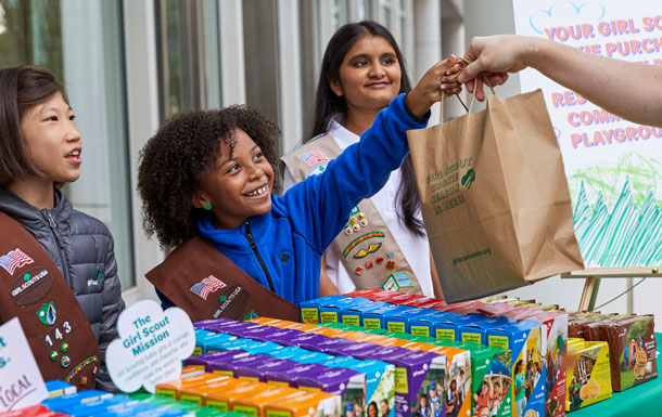 Three Girl Scouts stand behind a table of cookies and one hands a paper shopping back to the hand of a customer.