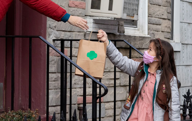 A Girl Scout wearing a pink face mask hands a brown paper bag with a green trefoil to someone standing on a porch. 