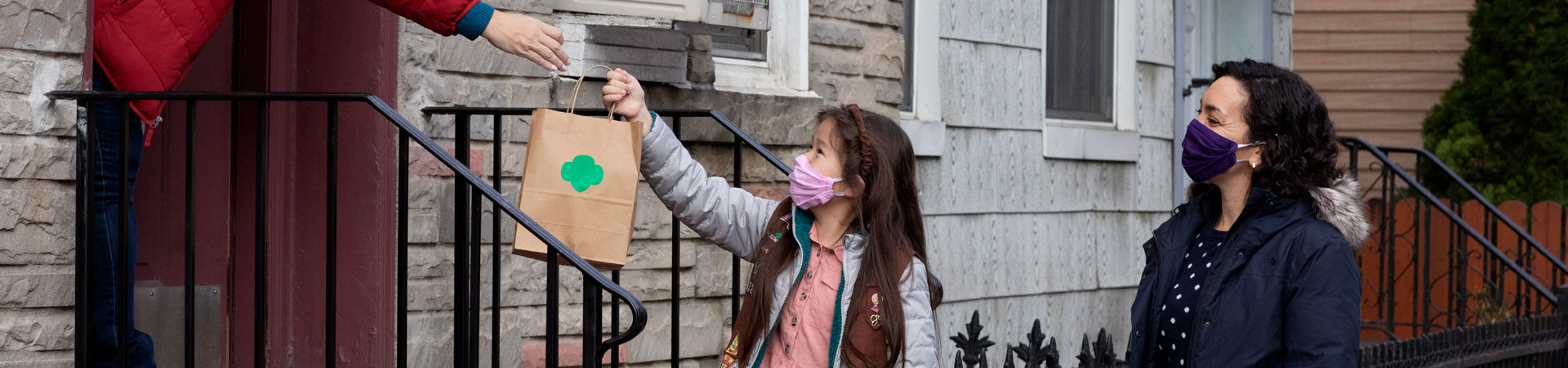  A Girl Scout wearing a pink face mask hands a brown paper bag with a green trefoil to someone standing on a porch. A masked caregiver supervises.  