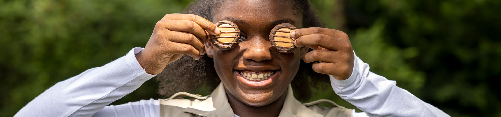  A close up of a Girl Scout wearing a khaki-colored vest holding two Adventurefuls Cookies in front of their eyes. 