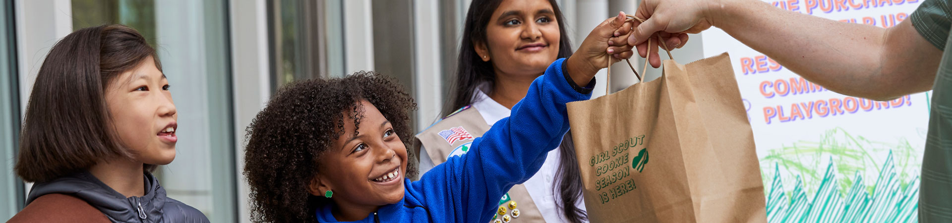  Three Girl Scouts at a Girl Scout Cookie booth. One is wearing a khaki vest decorated with pins and badges. Another hands a brown paper bag to a customer. The bag reads, "Girl Scout Cookie Season Is Here."  