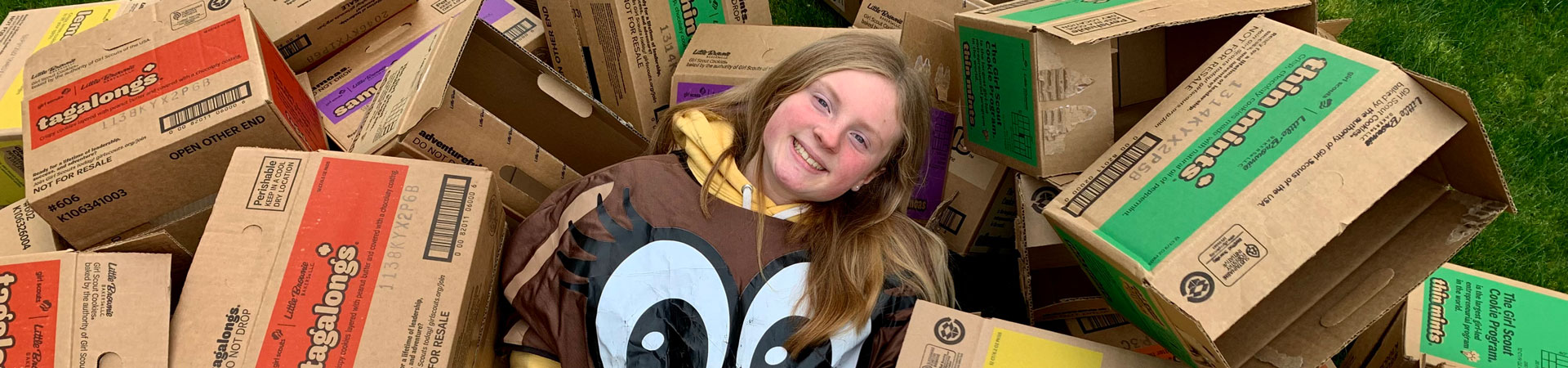  A Girl Scout dressed in a cookie costume surrounded by cases of various Girl Scout Cookies. 