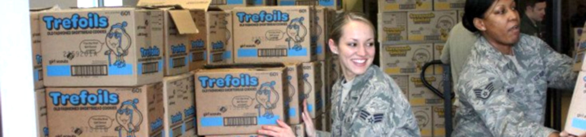  Two women dressed in military fatigues in a storeroom are surrounded by cases of Girl Scout Cookies. 