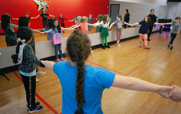 A group of youth Girl Scout members are in a gym with a woode floor and red walls. The Girl Scouts are standing around the perimeter of the room, arms extended, and holding each other's hands. 