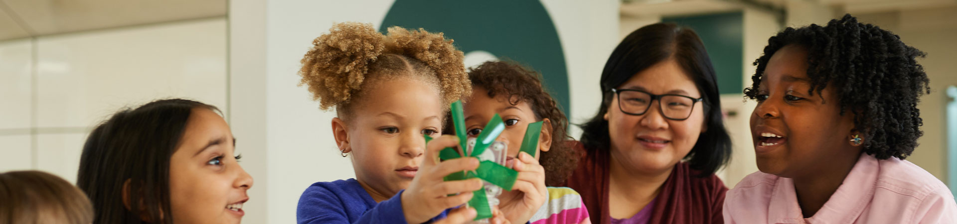  Four younger Girl Scouts are sitting at a table with an adult Girl Scout volunteer. There are sheets of paper on the table and they are working together to conduct  a science experiment using a plastic bottle. 