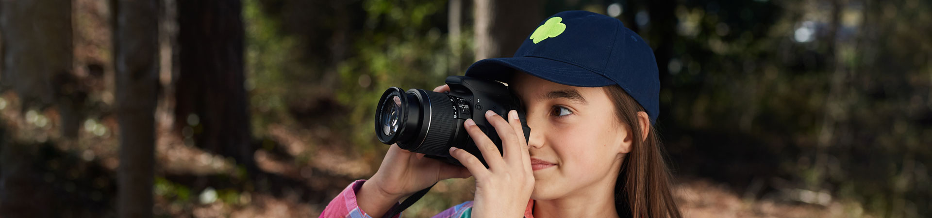  A Girl Scout wearing a blue baseball cap with a lime-green trefoil is standing outdoors looking through the viewfinder of a camera.  