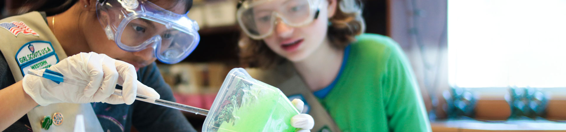  Two Girl Scouts are in a lab. They are wearing khaki-colored sashes embellished with pins and badges, safety goggles, and gloves. One Girl Scout is spooning a bright-green substance into a beaker. 