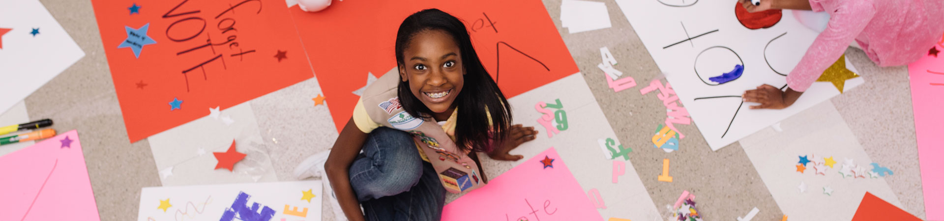  A Girl Scout wearing a khaki-colored vest decorated in various pins and badges is sitting on a floor scattered with various colored posters that read, "Vote."  
