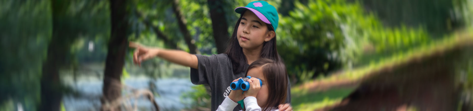  Two Girl Scouts stand outside in front of green trees and a body of water. The older Girl Scout wears a hat and points in the distance, while the younger Girl Scout looks through binoculars. 