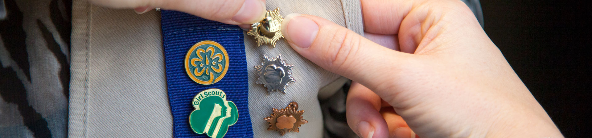 Two hands attaching a pin to a khaki-colored vest embellished with other Girl Scout pins. 