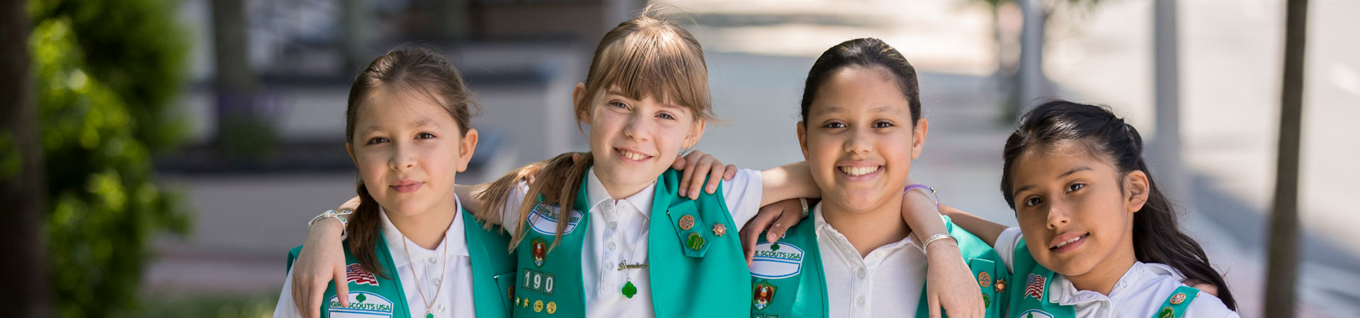  Four Girl Scouts wearing matching white shirts and green vests embellished with various pins and badges. They are standing outdoors with their arms around each other's shoulders.  