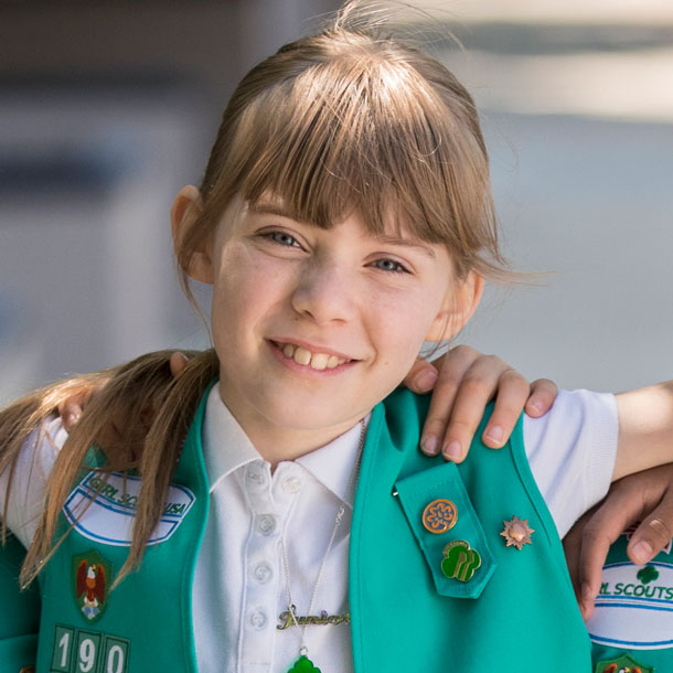 A Girl Scout wearing a white shirt and green vest embellished with a variety of colorful and metallic pins and other badges. 