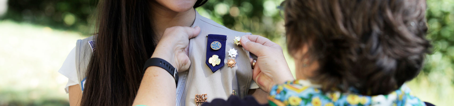  A Girl Scout wearing a khaki-colored vest embellished in a variety of metallic pins stands while an adult adds a gold pin to their vest. 