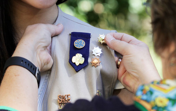 A Gold Award Girl Scout wearing a khaki vest decorated in pins and badges receives a Gold Award pin.