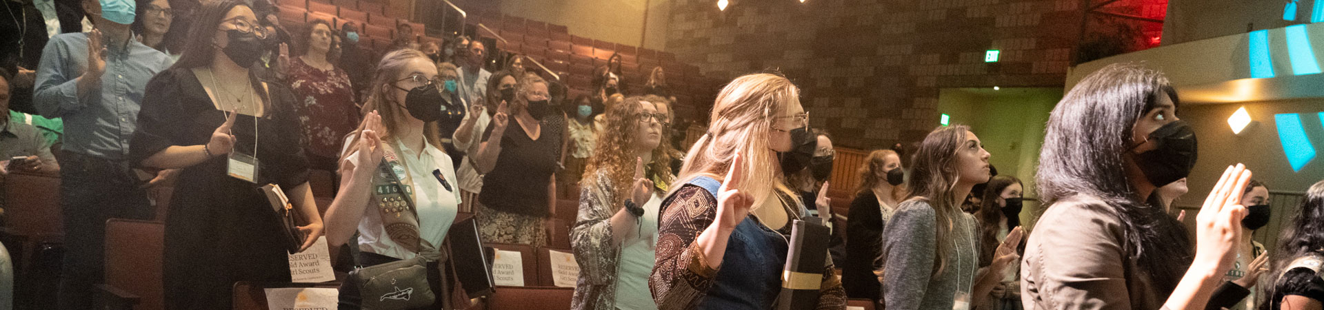  An auditorium is filled with masked Girl scouts. They are standing from their seats, looking to a stage, and making the Girl Scout sign with their hands.  