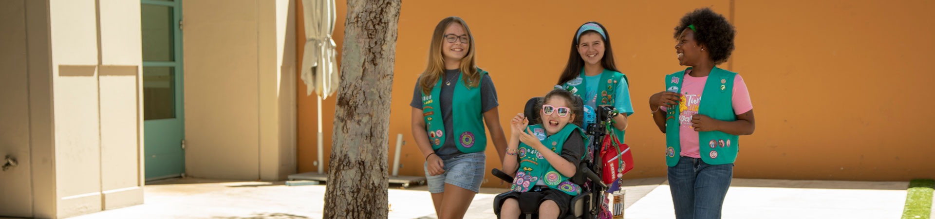  Four Girl Scouts wearing green vests decorated with various pins and badges. Three are walking and one is pushing the fourth in a wheelchair. They are on a paved path lined with walks. The sun is shining. There is a bright orange wall in the background behind them. 