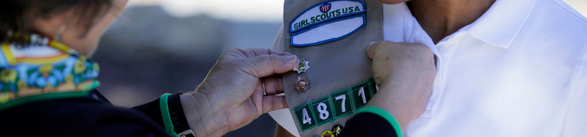  Two hands attach a silver pin to a Girl Scouts USA khaki-colored vest embellished with other metallic pins and badges.  