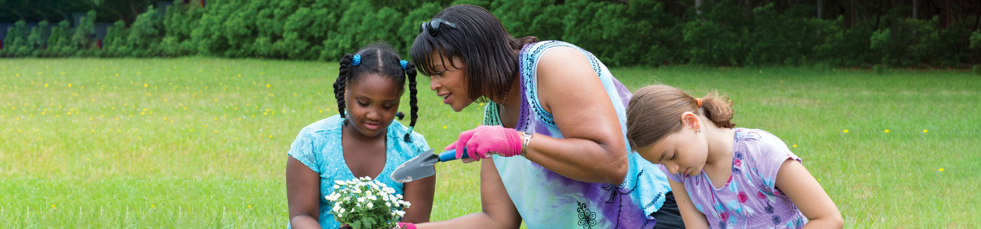  An adult volunteer and two youth Girl Scouts are sitting in the grass potting flowers. One of the youth Girl Scouts is holding a pot with white and green flowers. The adult volunteer is holding a small shovel.  