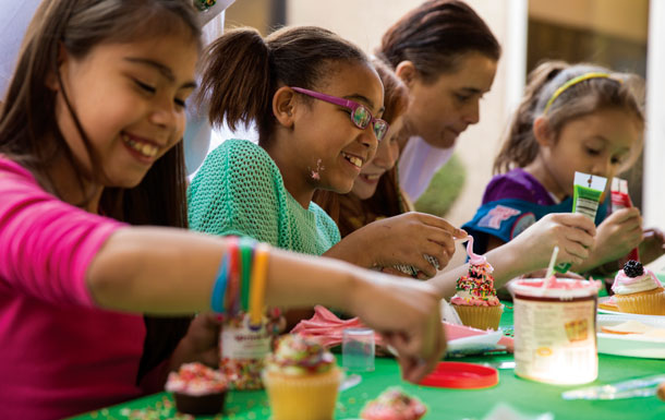 Girl Scouts sitting at a table decorating cupcakes while adult volunteers supervise.