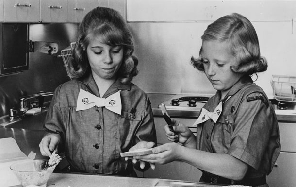 Image Description: Two Girl Scouts wearing buttoned t-shirts and bow ties make a sandwich together in the kitchen.