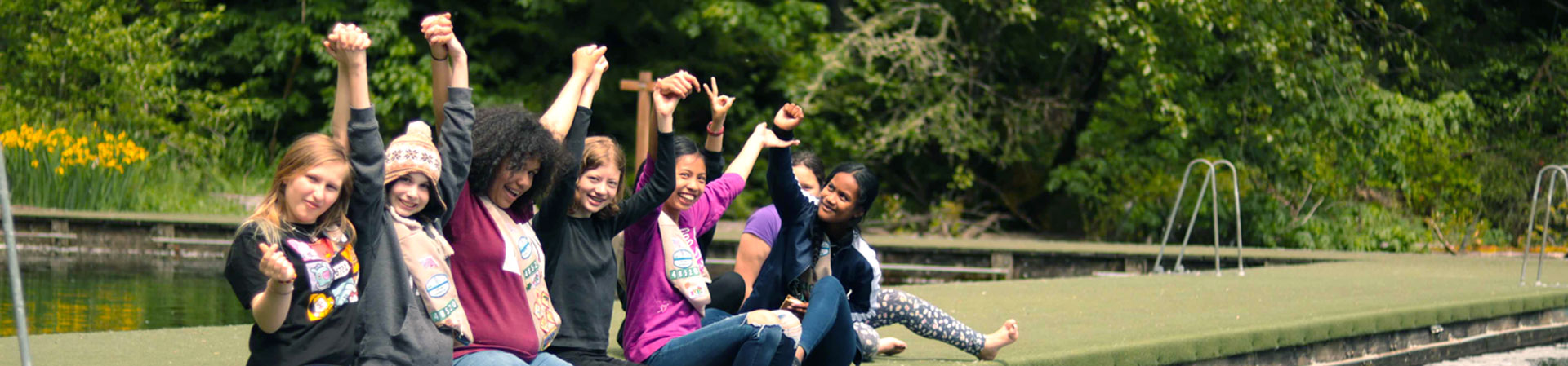  Image Description: A group of Girl Scouts sits on a dock on a lake. They are in a row with their arms raised high over their heads, and they are holding each other's hands.  