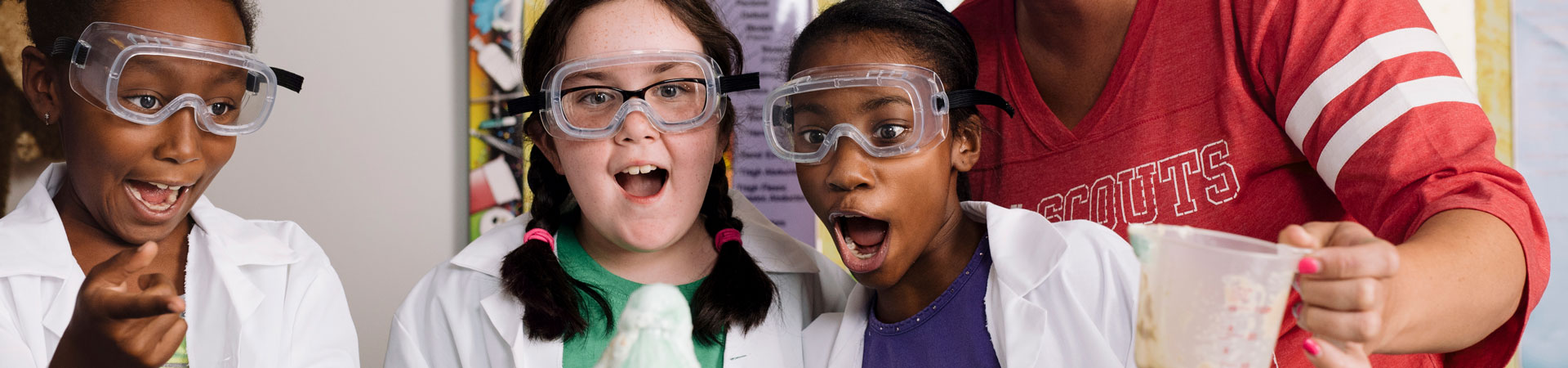  Three Girl Scouts wearing lab coats and goggles. They appear amazed at the results of a science experiment. 