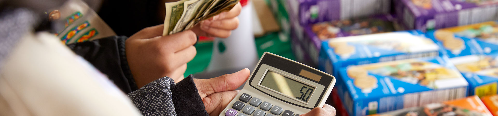  A person's hands are holding a calculator while the hands of another person counts money. There is a table stacked with boxes of Girl Scout Cookies in the background. 