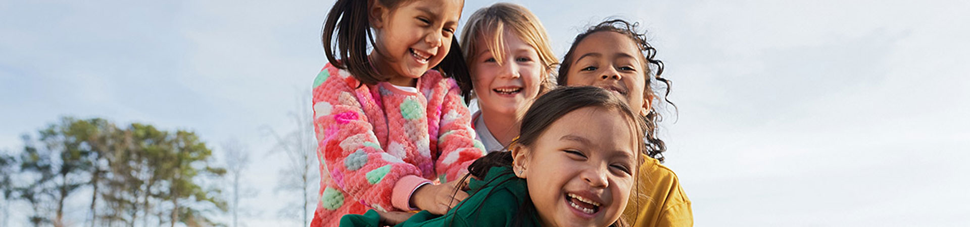  Four younger Girl Scouts playing in the grass. One Girl Scout is laying in the grass laughing. Three others surround her smiling. The sky is blue above them. 