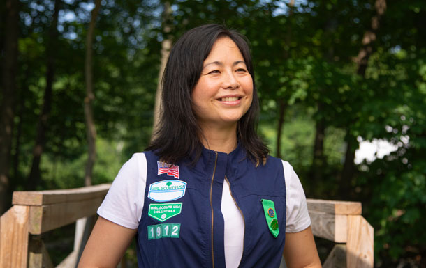 An adult volunteer wearing a white shirt and blue vest decorated in various badges stands outside with trees behind them. 