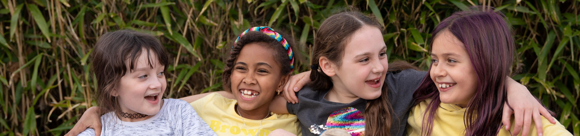  Four Girl Scouts are sitting on the ground. They are smiling and have their arms around each other's shoulders. There is tall grass behind them.  