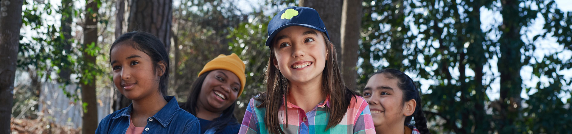  Four smiling Girl Scouts walk through the woods. They are wearing colorful Girl Scout apparel and there are trees behind them. 
