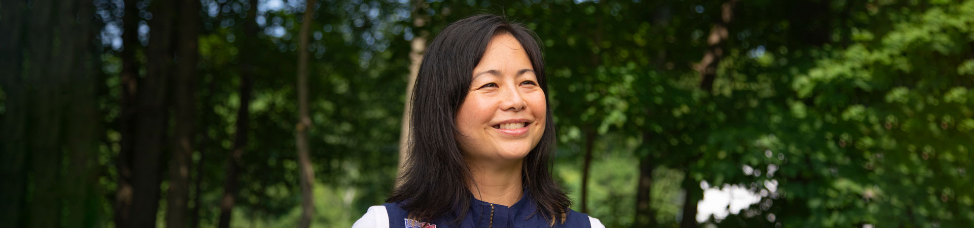  Adult woman girl scout volunteer wearing a blue vest stands outdoors. There are trees in the background. 