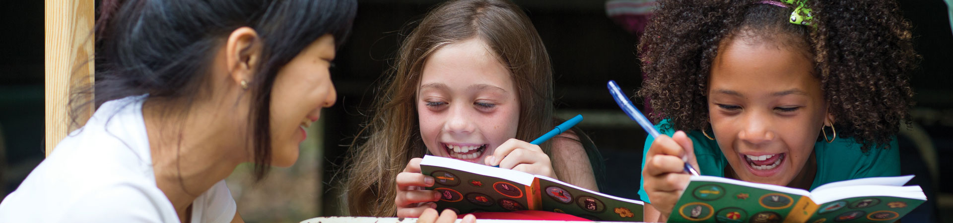  An adult volunteer and two younger Girl Scouts are gathered. The two youth Girl Scouts are smiling and writing in notebooks. 