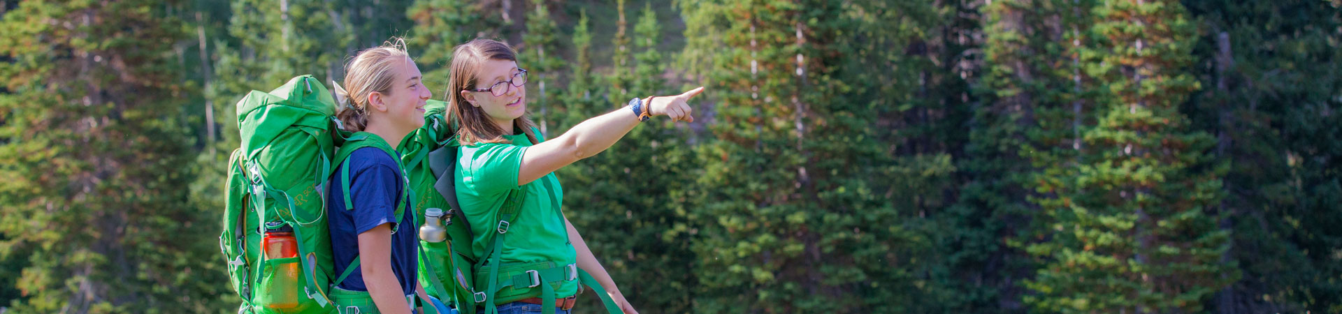  Two Girl Scout volunteers wearing green backpacks are hiking. One is pointing to something off in the distance. There are trees behind them. 
