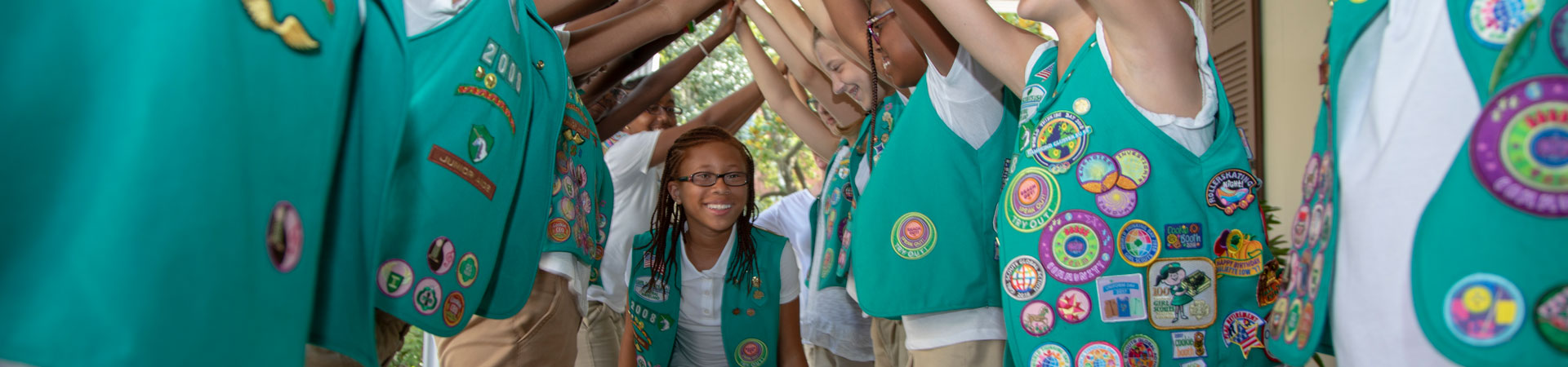  A group of Girl Scouts dressed in khaki shorts and green-colored vests embellished in various pins and badges raise their arms to create a tunnel. One Girl Scout walks through the tunnel. 