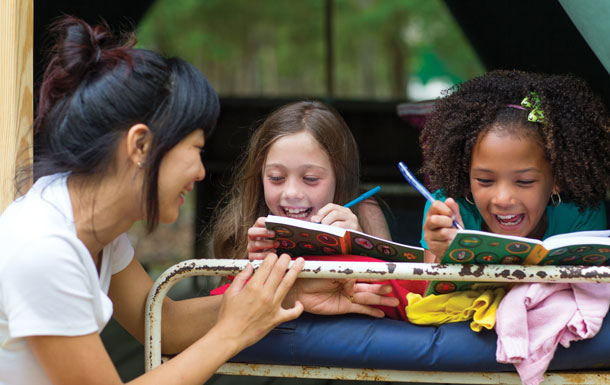 A smiling adult Girl Scout volunteer and two smiling youth Girl Scout members. The youth members are writing in notebooks.