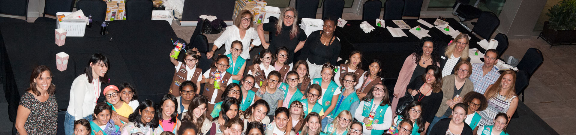  A group of adult Girl Scout volunteers and youth Girl Scout members are gathered for a service unit meeting. Many are wearing green or brown vests embellished in a variety of pins and badges.  